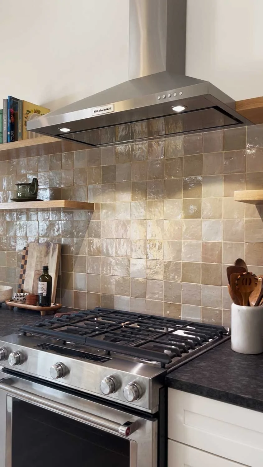 Stainless steel range hood over a kitchen stove with a beige tiled backsplash and wooden floating shelves holding books and kitchen utensils.