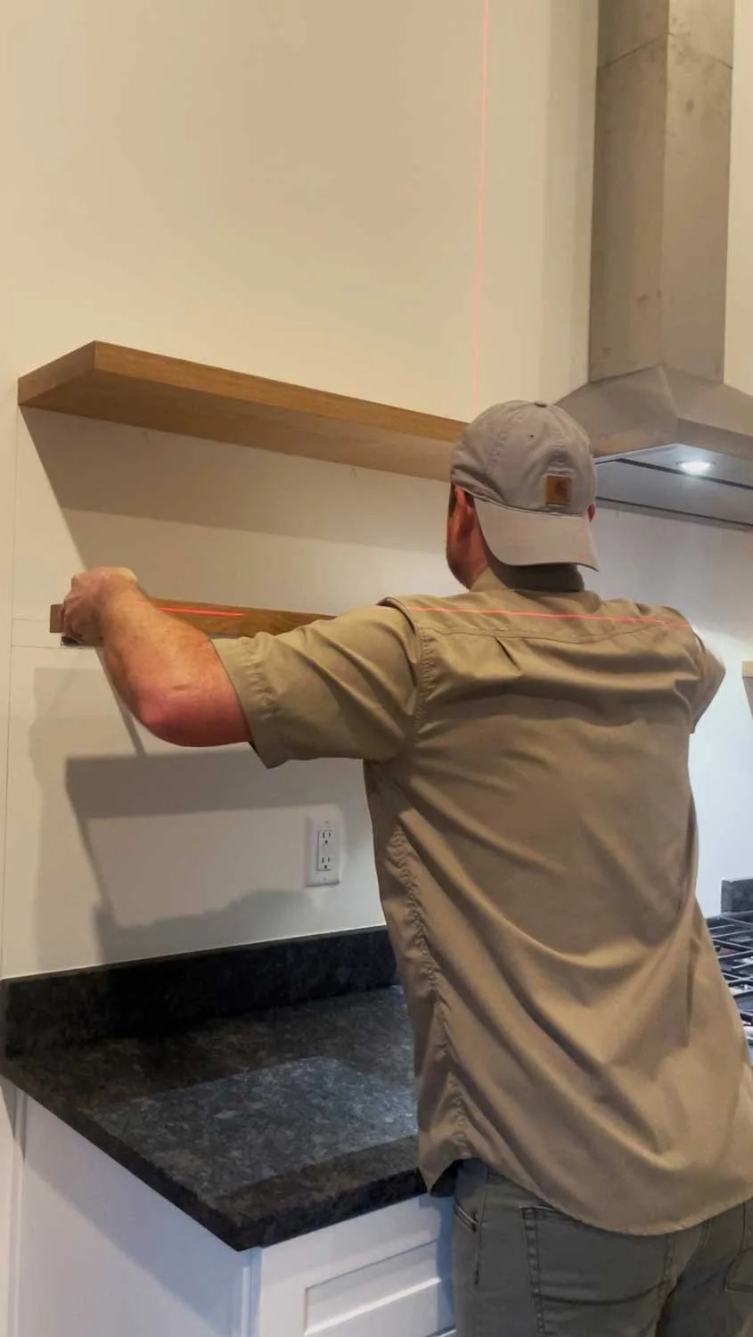 A man installing a wooden floating shelf on a kitchen wall, using a laser level to ensure it is straight.