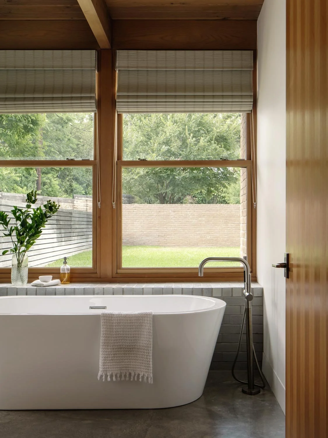 Modern bathroom with a white freestanding bathtub and large windows showing a green backyard beyond.