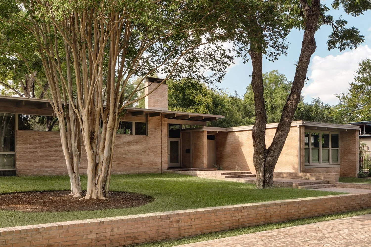 Mid-century modern brick house with flat roof, large windows, surrounded by green lawn and mature trees.