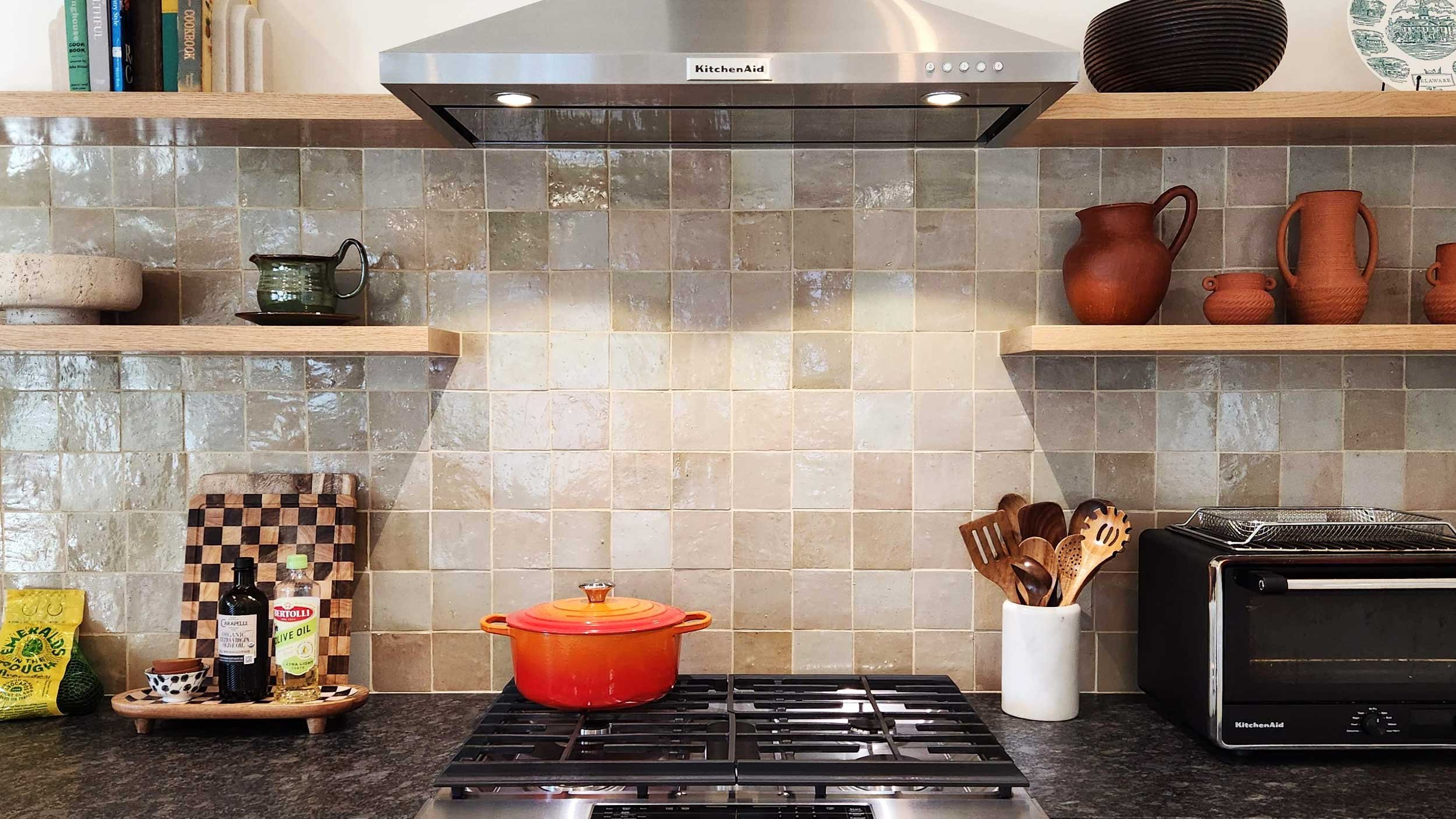 Kitchen backsplash with beige tile, wooden floating shelves holding books and decorative pottery, and a stainless steel vent hood.