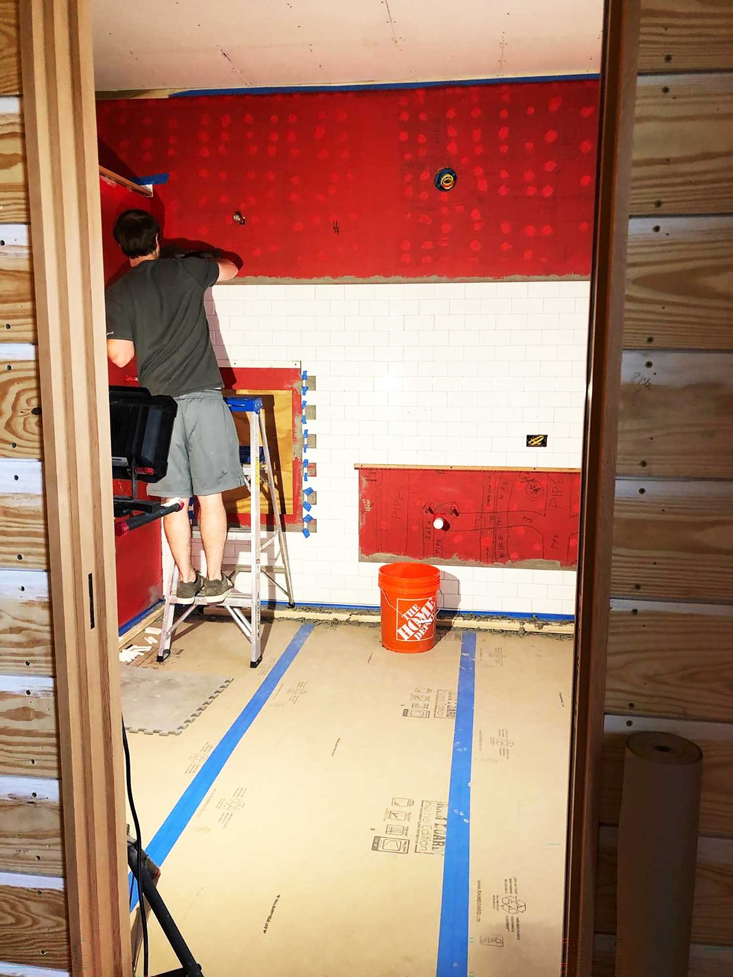 Man standing on a ladder installing white subway tile on a bathroom wall.