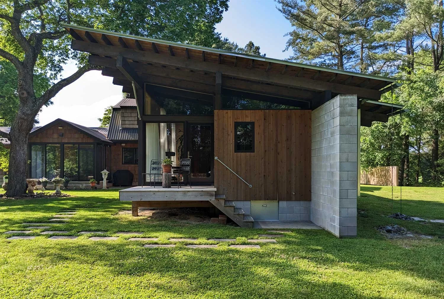 Modern house with a wooden and concrete exterior, a small elevated deck with a table and chairs, surrounded by a green lawn and trees.