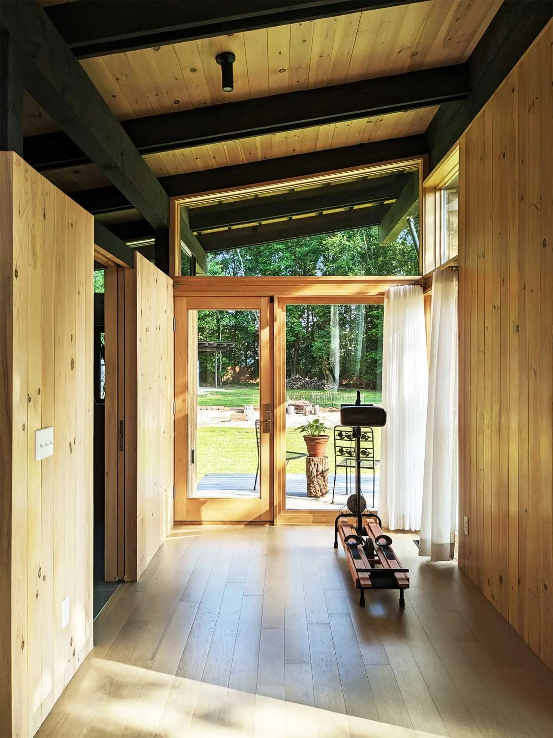Sunny interior of a home with wooden walls and ceiling, glass door leading outside, and an exercise machine in the room.