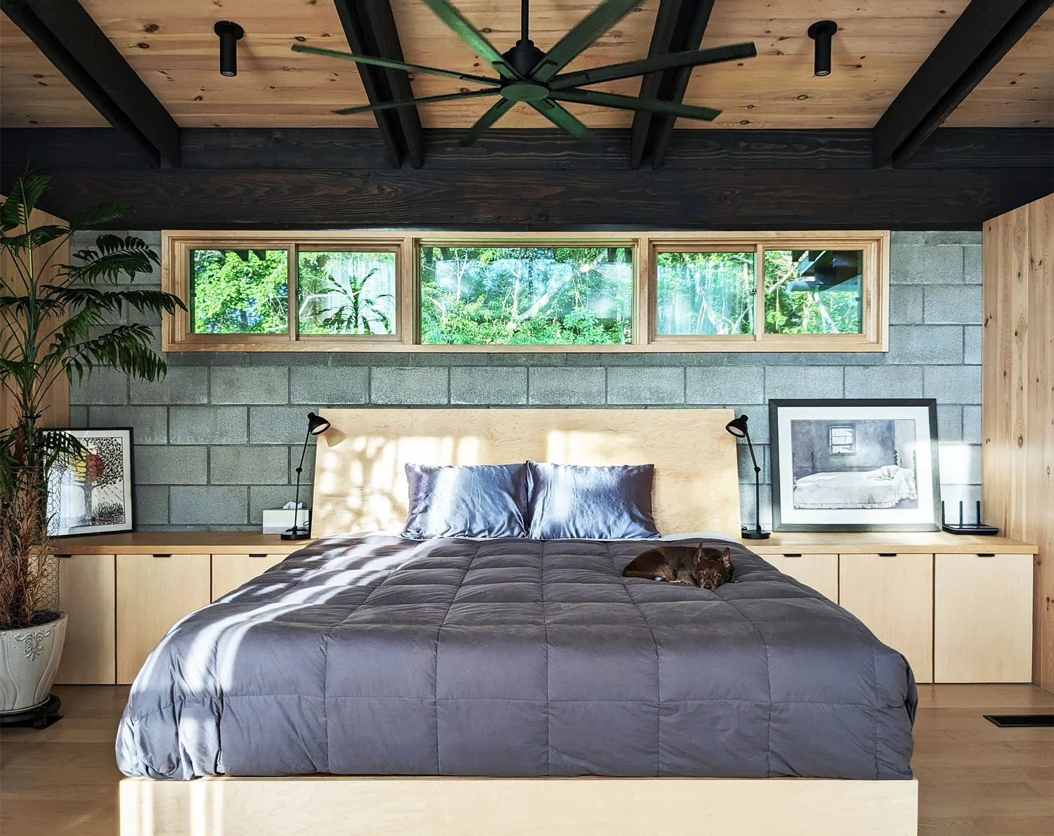 Modern bedroom with wooden built-in bed and cabinets, and greenery outside visible through clerestory windows.