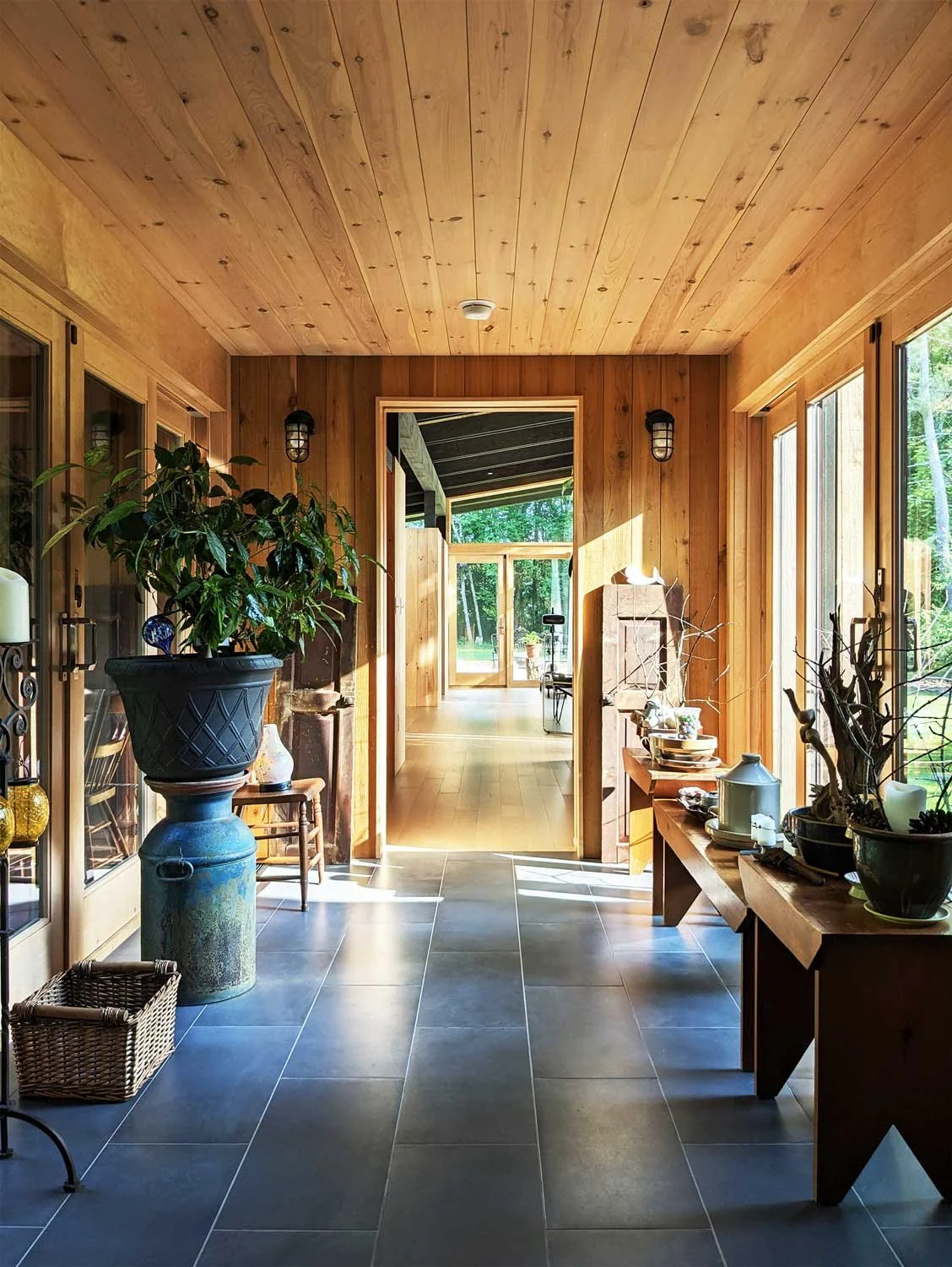 View of a sunlit wooden breezeway with large windows, decorated with potted plants, leading to a modern bedroom wing.