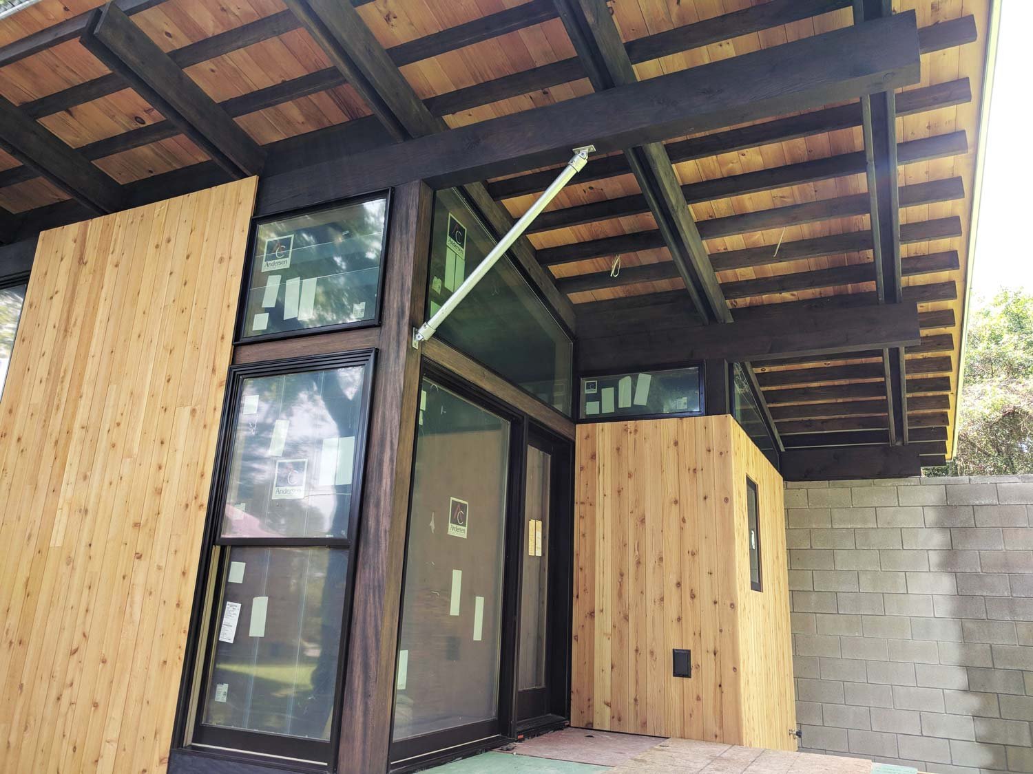 Exterior of a modern house under construction with wood and glass siding, with large windows, a covered porch, and a concrete block wall.