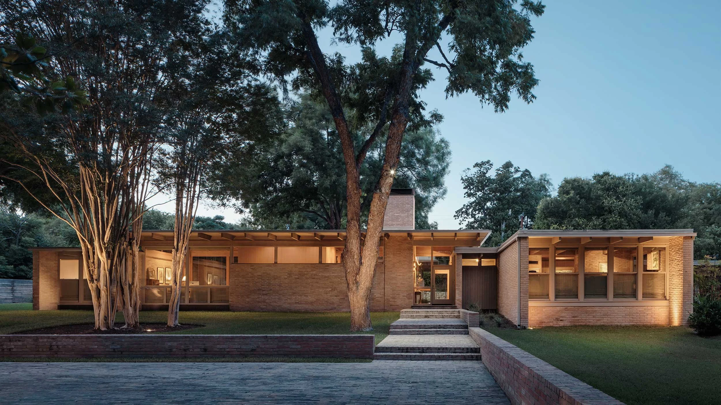 Modern brick house with large windows and a flat roof, illuminated from inside, surrounded by trees in the evening.