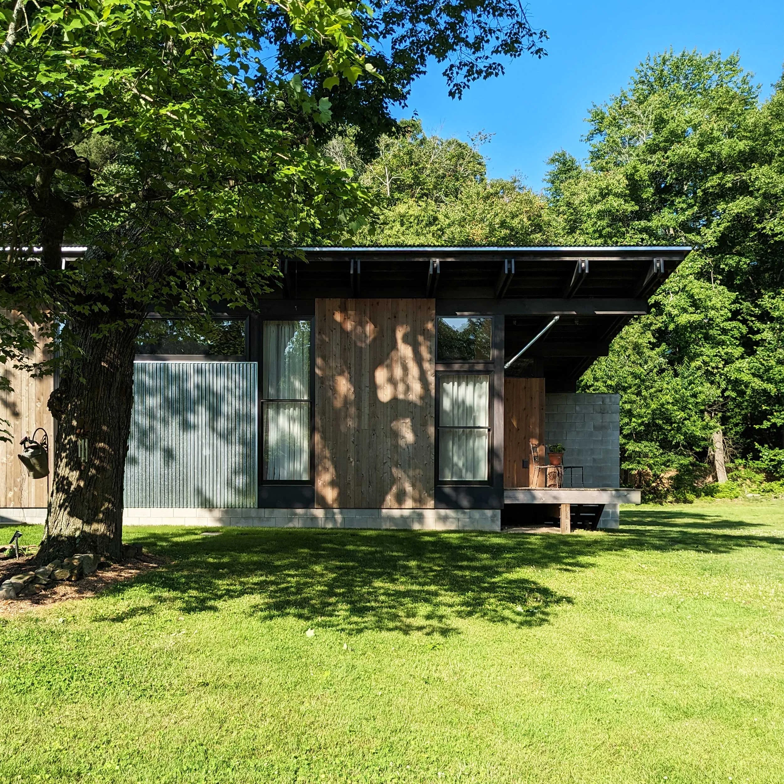 A modern house with large windows and wooden siding surrounded by a garden with green grass, a large leafy tree on the left, under a clear blue sky.