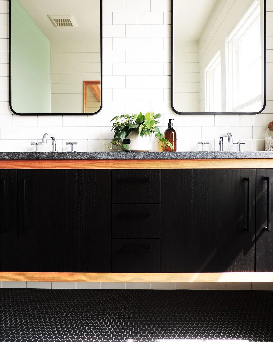 Bathroom vanity with two mirrors, a plant, a soap dispenser, black cabinet, and tiled wall.