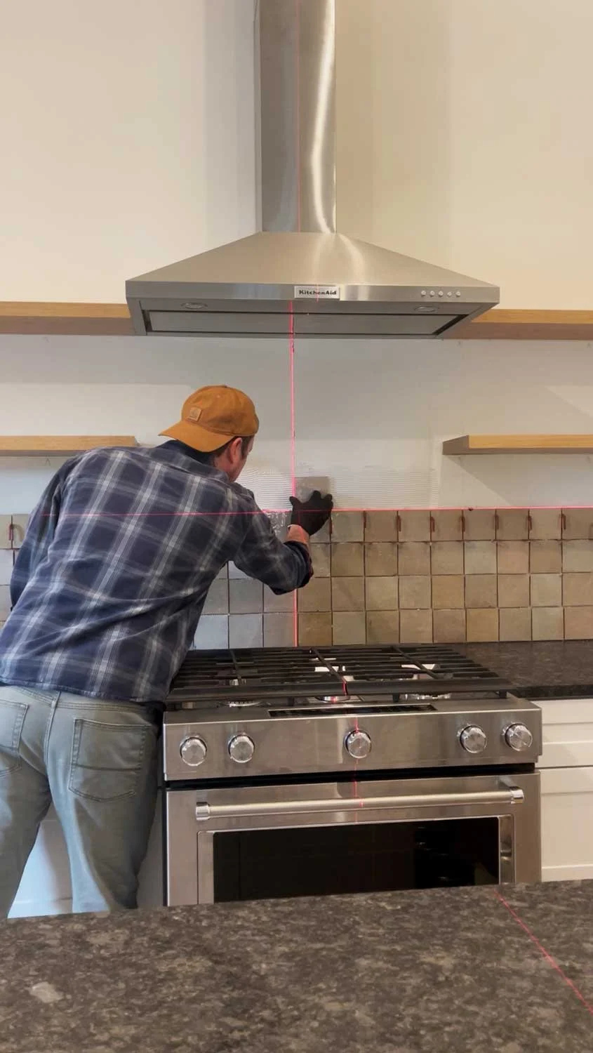 A man installing kitchen backsplash tile using a laser level to ensure it is level while standing in front of a stove, with a kitchen range hood above.