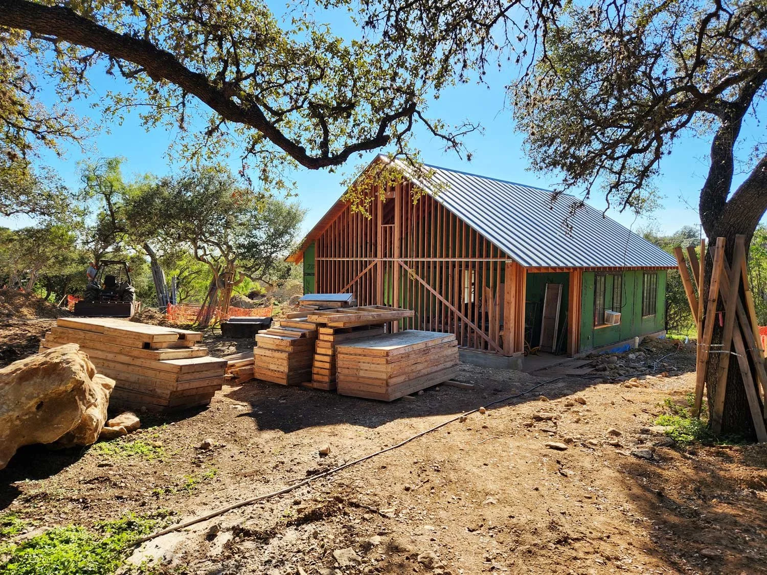 Small house under construction with wood framing, stacks of lumber, large rocks, and construction equipment.