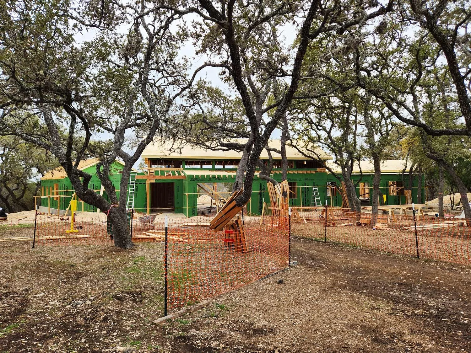 Construction site of a house with green sheathing, surrounded by trees, with orange safety netting and construction materials.