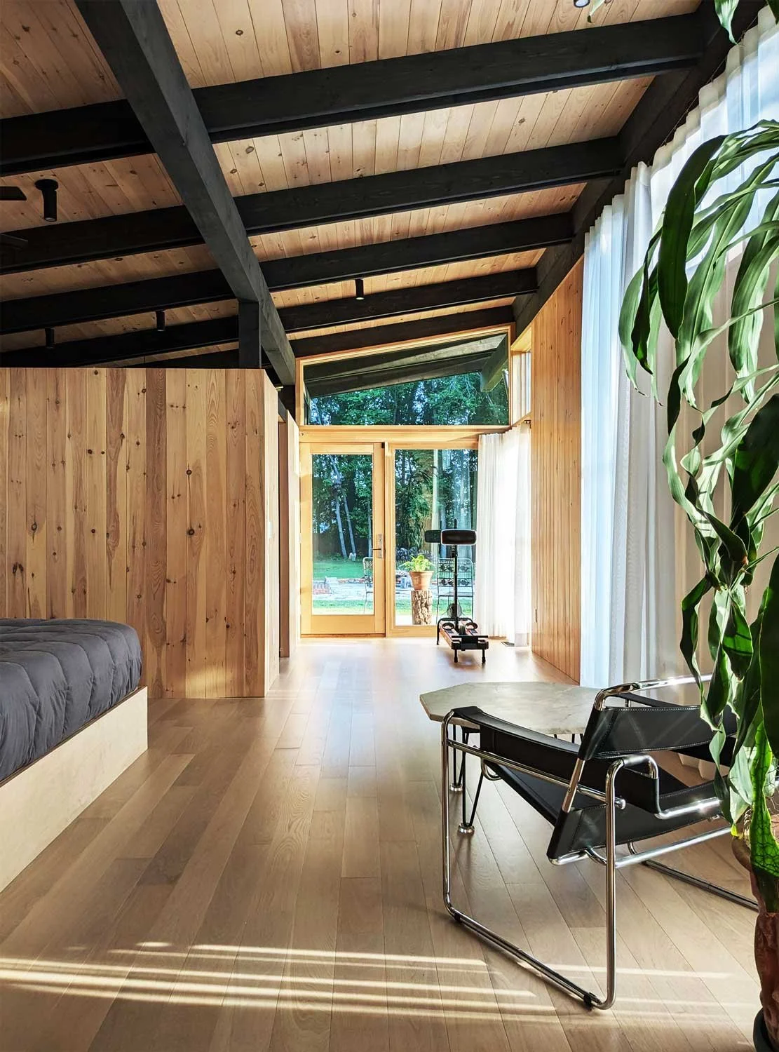 Bright bedroom with wooden walls, exposed ceiling beams, and large window and glass door leading outside.
