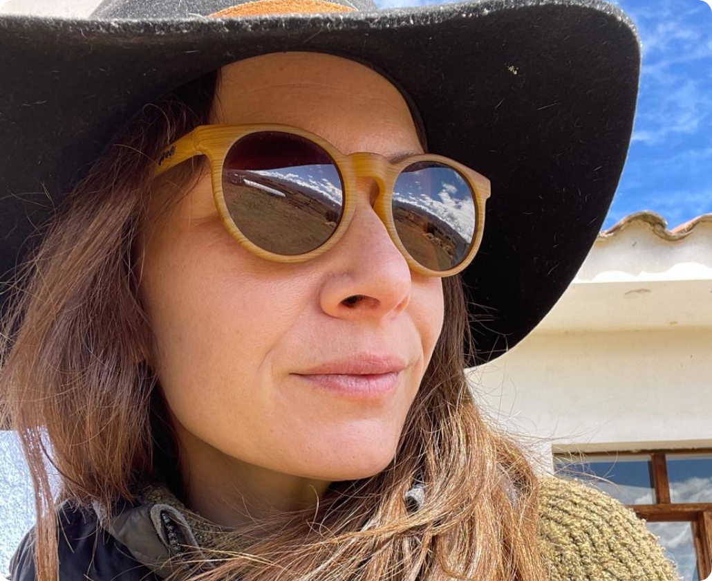 Close-up of a woman wearing a large black hat and round wooden sunglasses, with a reflection of a blue sky and clouds in the sunglasses, outdoors.