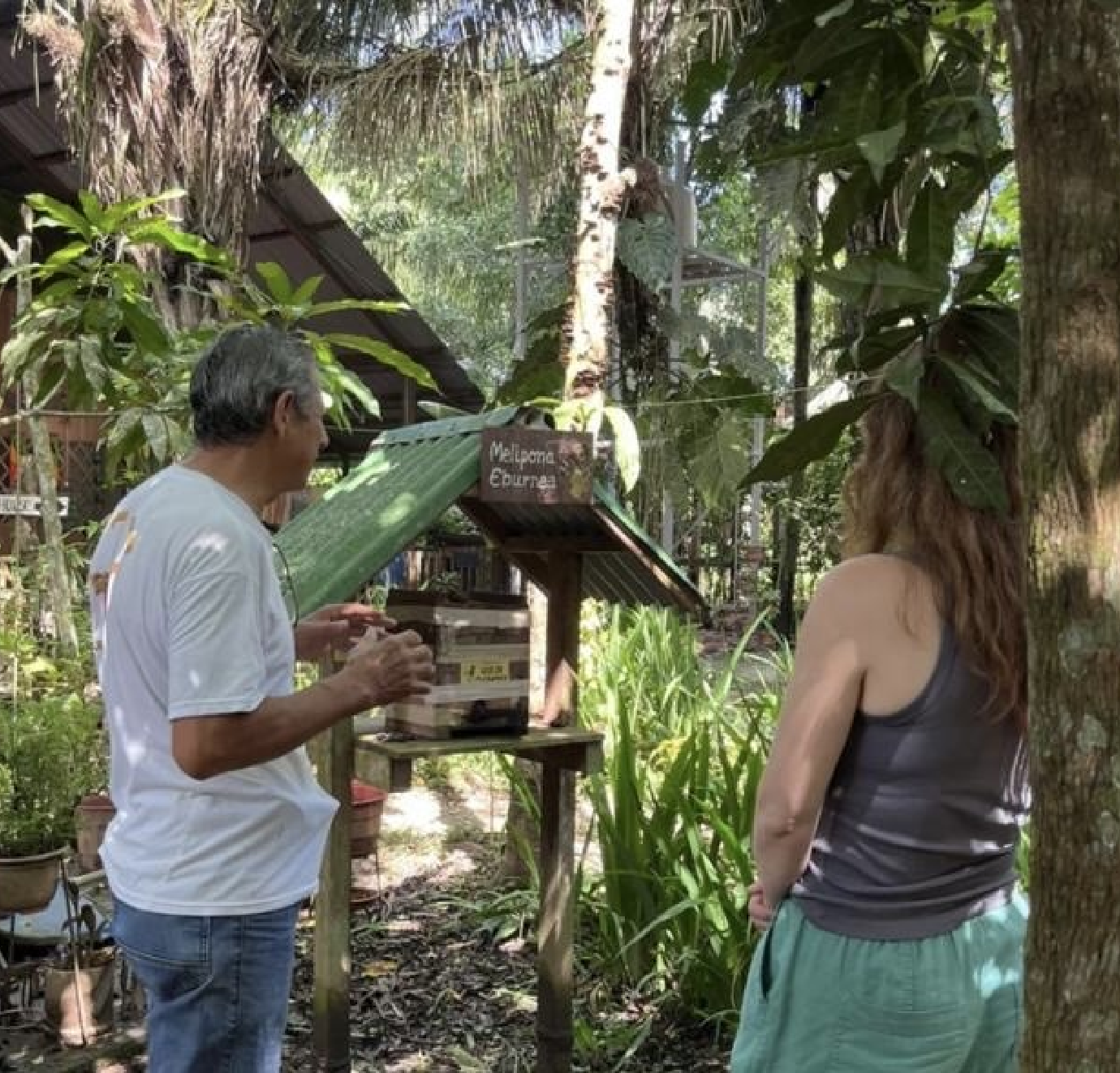A man and woman standing outdoors in a lush, green tropical setting, looking at a sign that reads 'Malpura Sanctuary' or similar. The man is wearing a white shirt and jeans, the woman has long hair and is dressed in a sleeveless top and green pants, with trees and plants surrounding them.