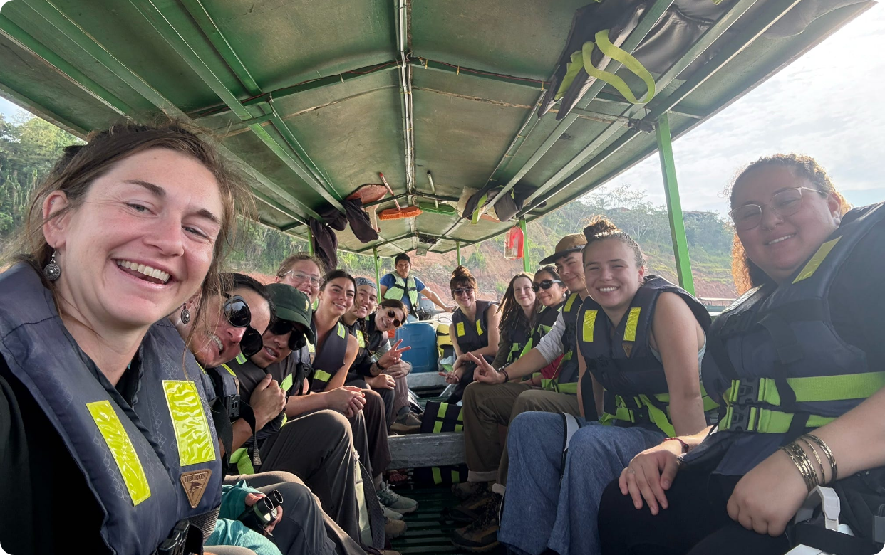 A group of people sitting inside a boat with green covering, all wearing life jackets, smiling for the camera during daytime.