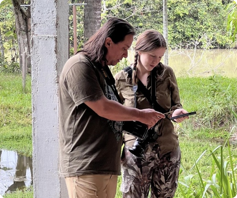 Two people near a pond or lake, looking at a phone together, one with a camera strap around their neck, surrounded by greenery.