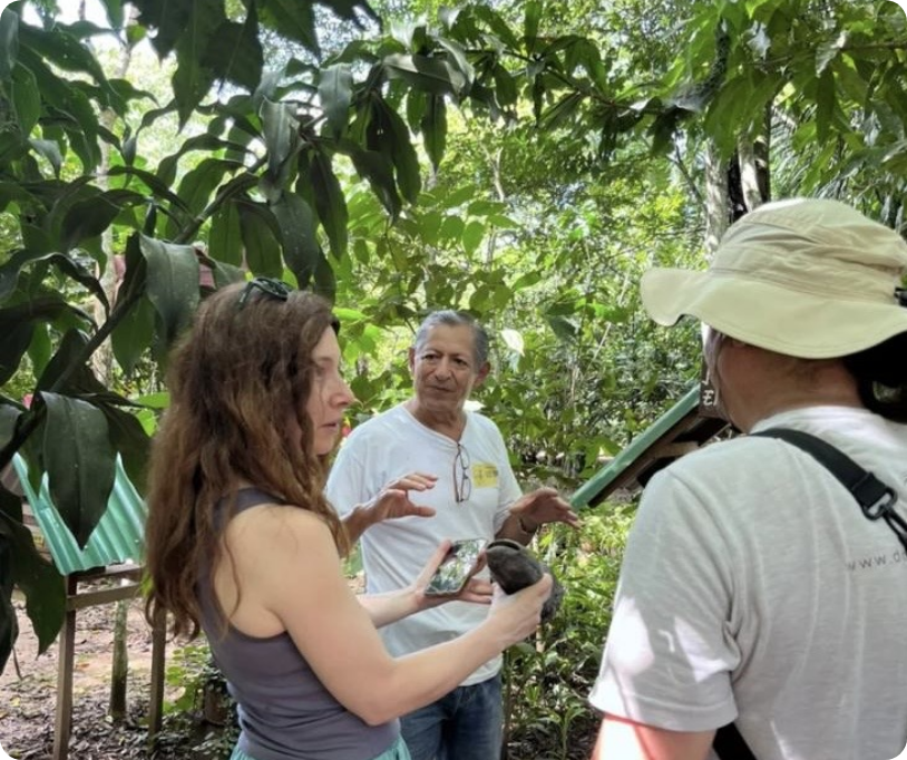 Three people are in a lush green jungle setting, engaging in a conversation. One woman is holding a small animal and a smartphone, while a man in a white shirt is gesturing with his hands. Another person with a backpack and wide-brimmed hat faces away from the camera.