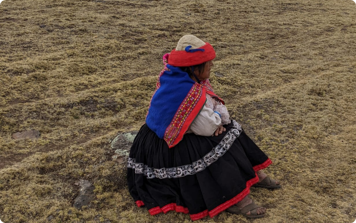A woman sitting on a small rock in a dry, grassy landscape, wearing traditional Peruvian clothing with a colorful shawl and head covering.