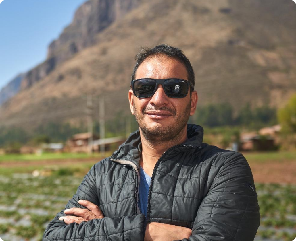 A man wearing sunglasses and a black jacket with crossed arms outdoors, with a mountain and field in the background.