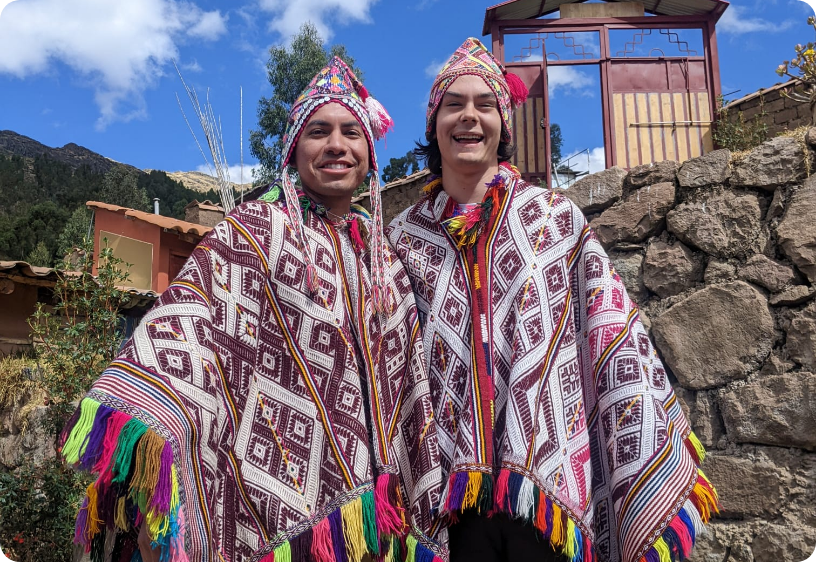 Two people wearing traditional indigenous clothing with intricate diamond patterns, colorful tassels on the edges, and headdresses, standing outdoors near a stone wall and a small building under a blue sky.