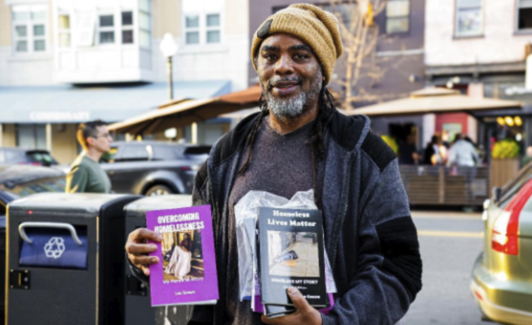 Leo Gnawa holding two books that he self-published as a homless man, writing about his experiences with homelessness, in Homeless Lives Matter and Overcoming Homelessness.