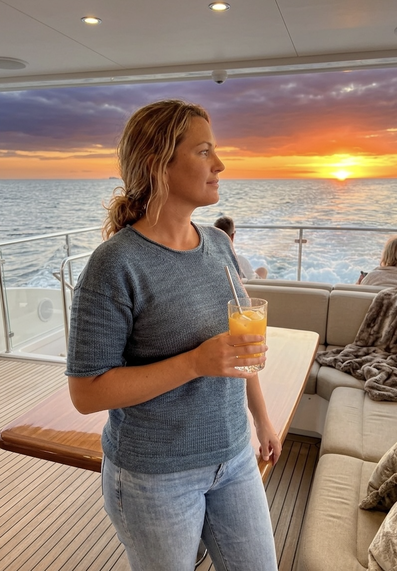 A woman with wavy blonde hair wearing a gray shirt and blue jeans, holding a glass of orange juice with a straw, stands on a boat deck during sunset with the ocean in the background.