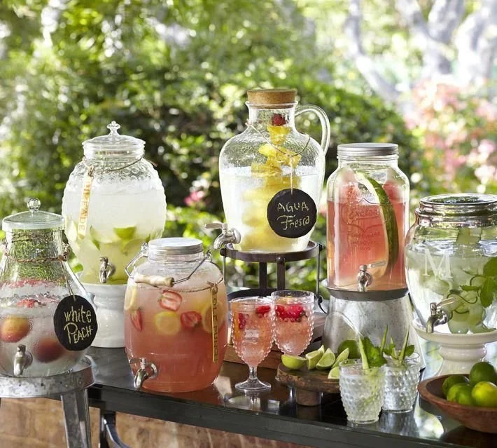 Glass beverage dispensers filled with infused water and fresh fruit, labeled with chalk signs, placed on a table outdoors.