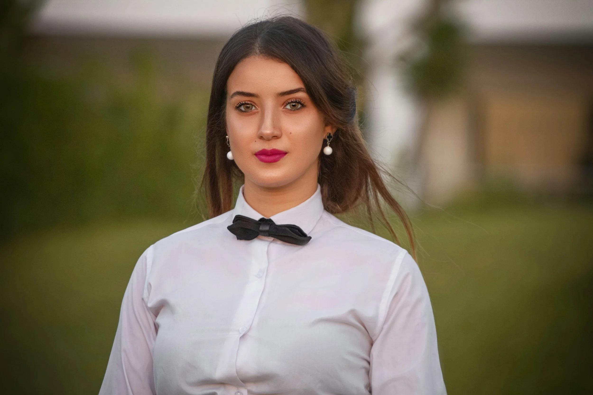 A young woman with dark brown hair, wearing a white shirt with a black bow tie, pearl earrings, and pink lipstick, standing outdoors with blurred greenery in the background.