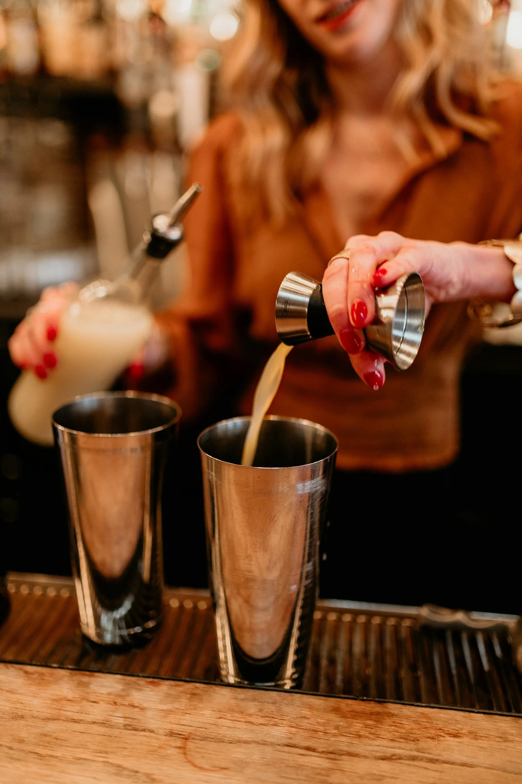 A woman with red nails is pouring a drink into two metal cups at a bar, with one hand holding a shaker and the other pouring from a bottle.