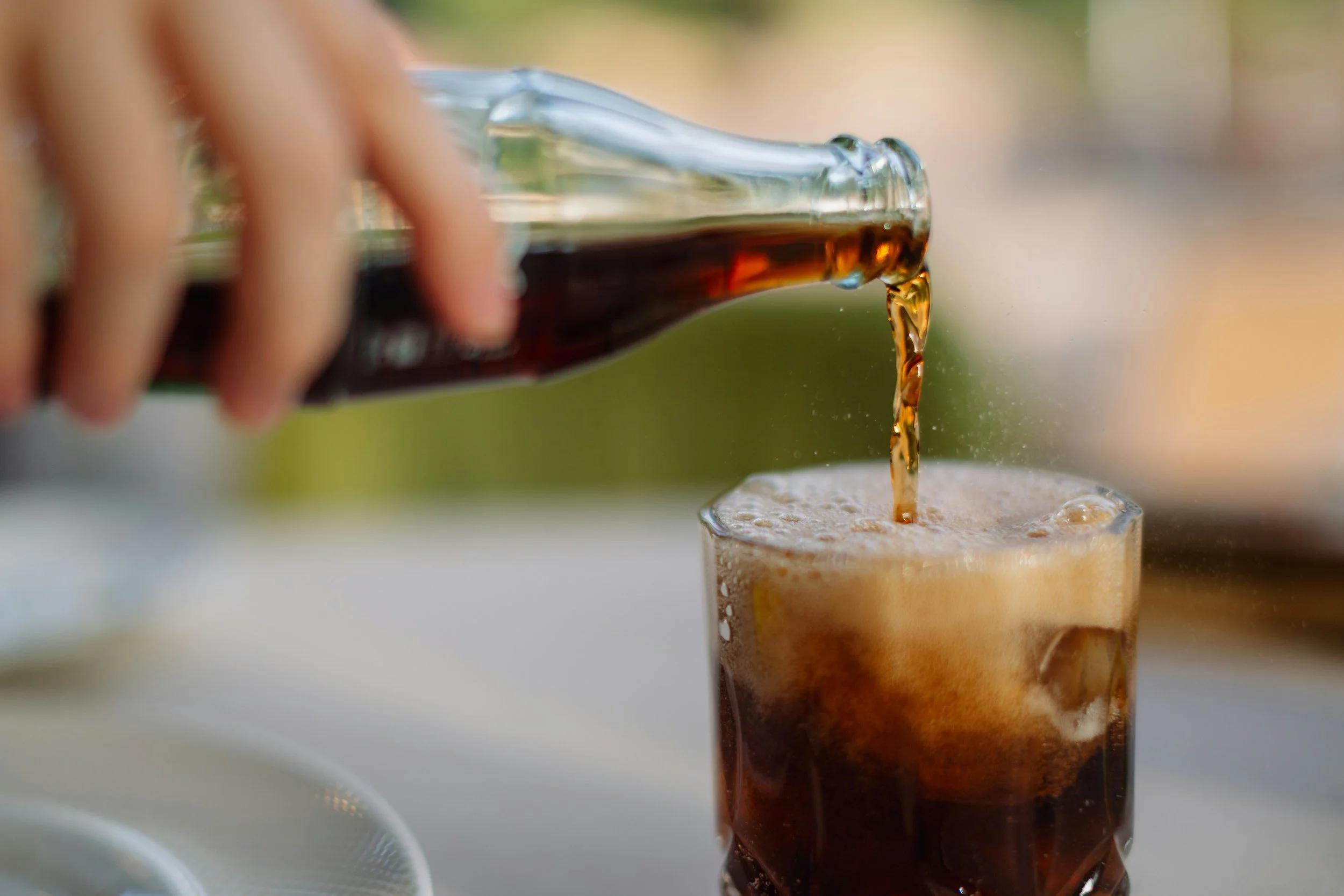 A hand pouring cola soda from a glass bottle into a glass filled with ice, creating foam on top.