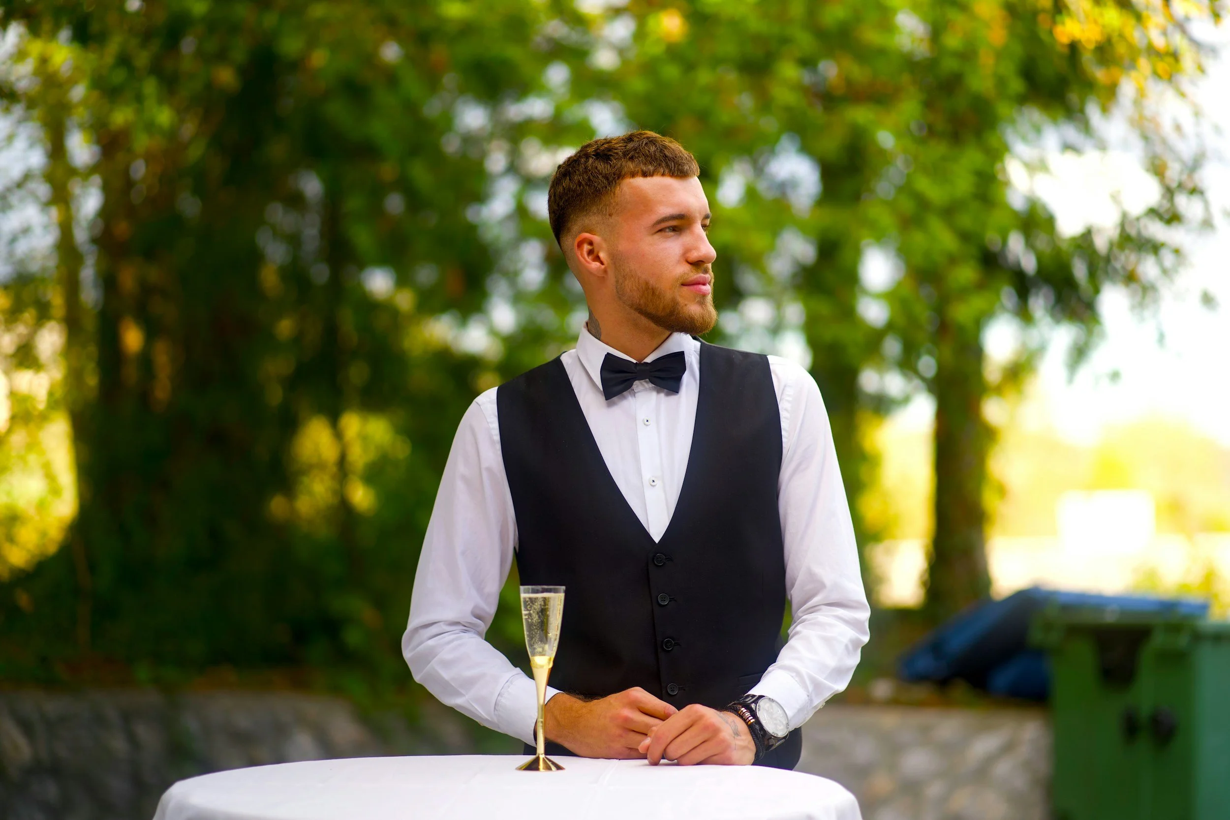 Young man in formal attire with white shirt, black vest, and bow tie standing outdoors with a glass of champagne on table, green trees in background.