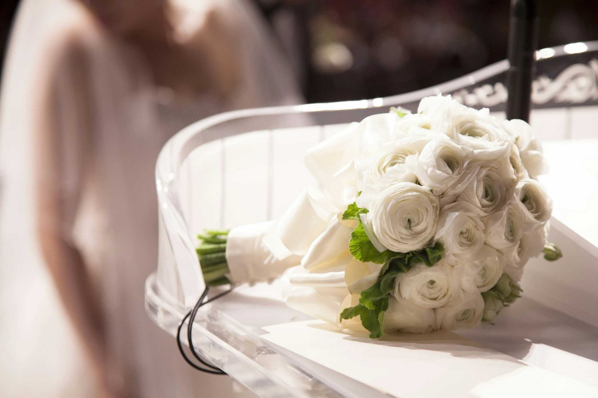 A bouquet of white roses and ranunculus flowers with green leaves, resting on a white surface, with a blurred background.