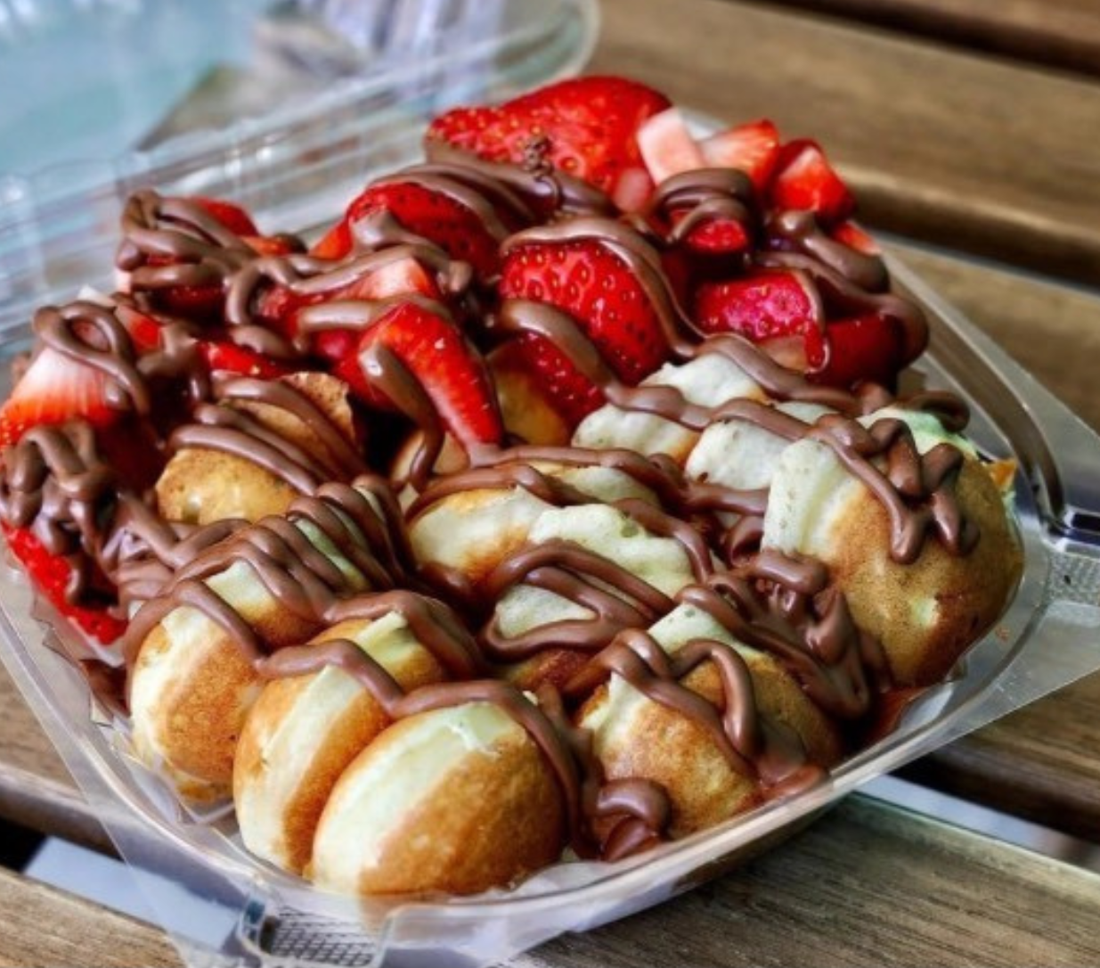 A dessert with mini pancakes topped with sliced strawberries, drizzled with chocolate sauce, in a plastic container on a wooden surface.