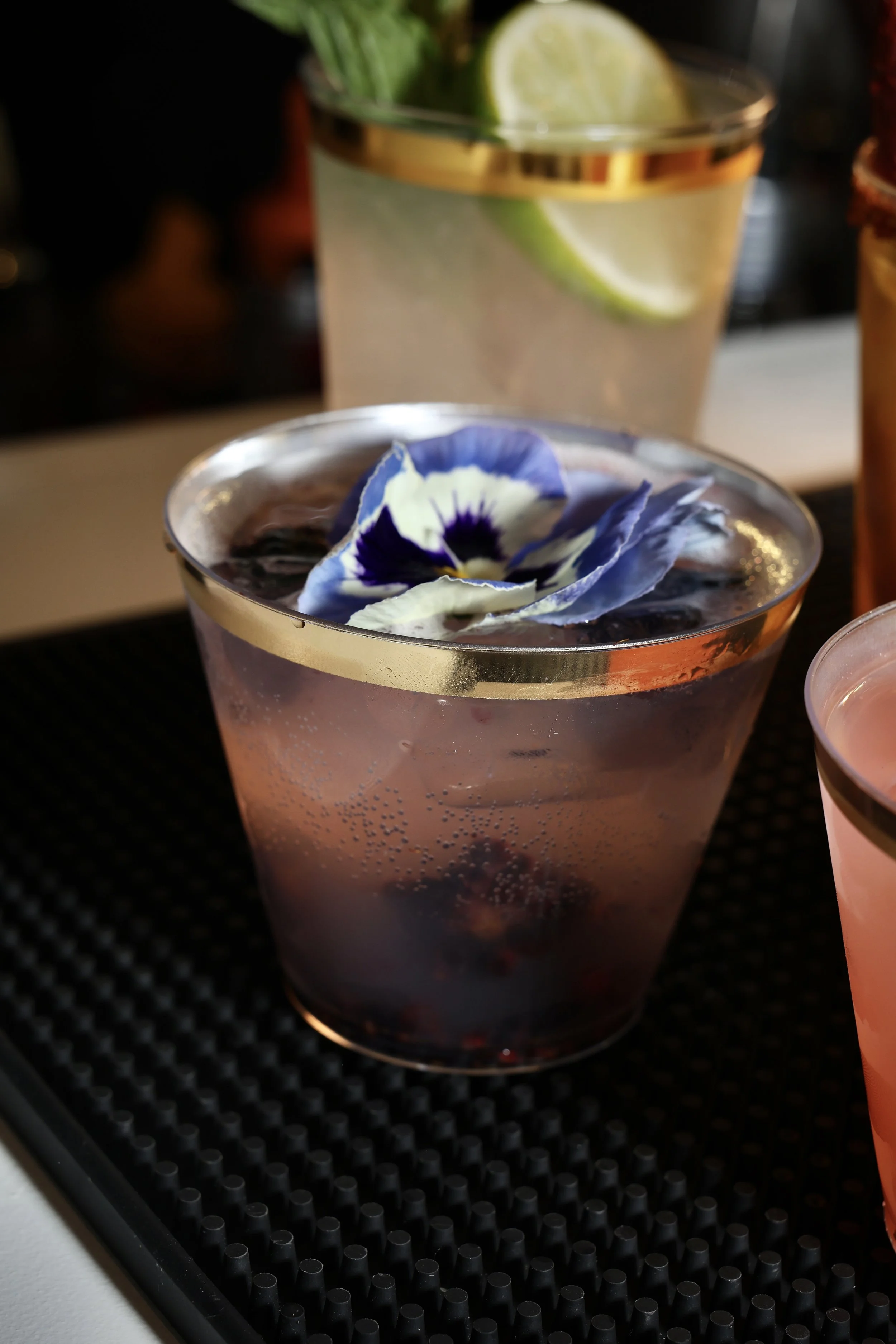 A cocktail drink in a glass with a gold rim, garnished with an edible purple and white flower, placed on a bar mat.