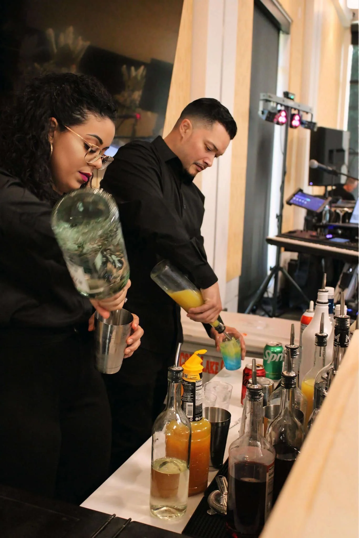 Two bartenders, a woman with glasses and a man, preparing colorful cocktails behind a bar with various bottles and mixers.