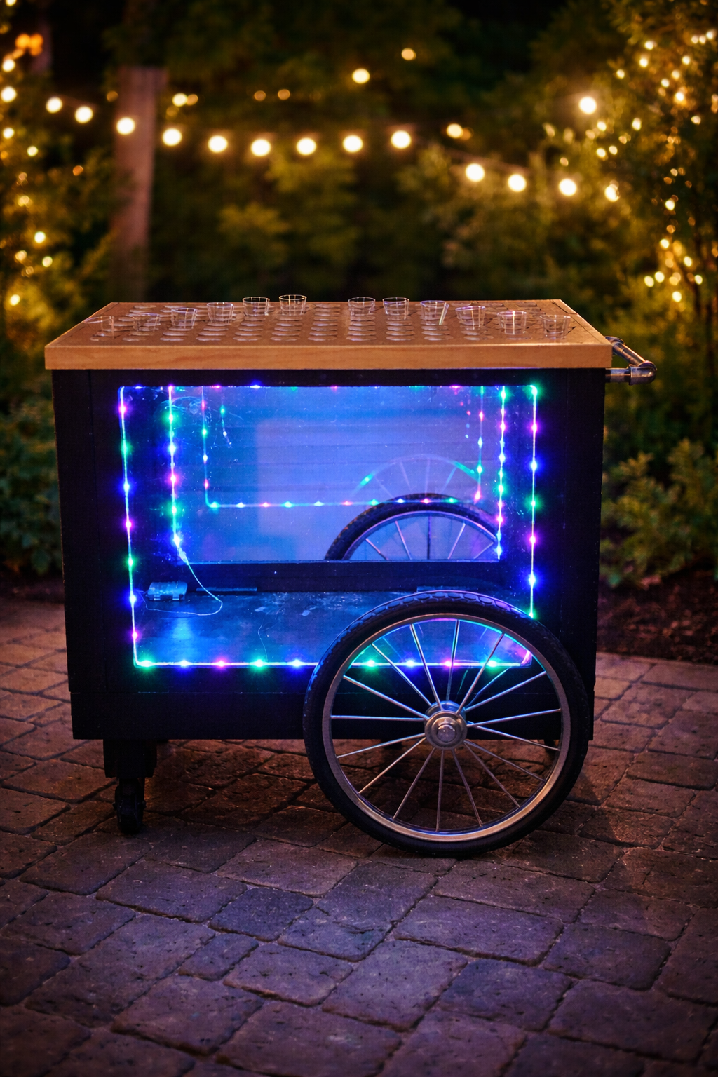 A lit-up carnival-style popcorn machine with a glass window, two large wheels, and a wooden top, decorated with colorful LED lights, set outdoors on a brick surface at night.