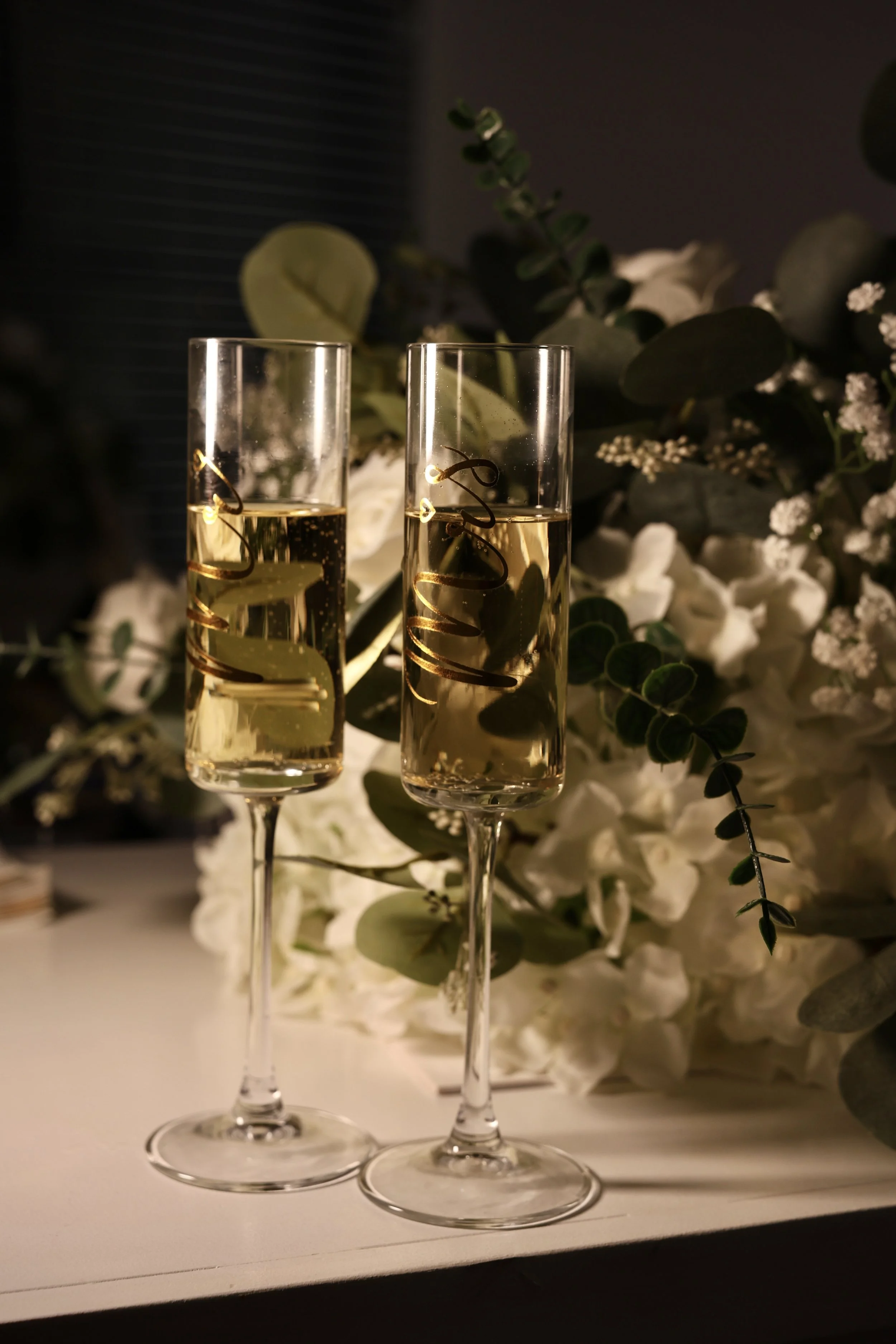Two champagne flutes filled with champagne, set against a backdrop of white flowers and greenery.