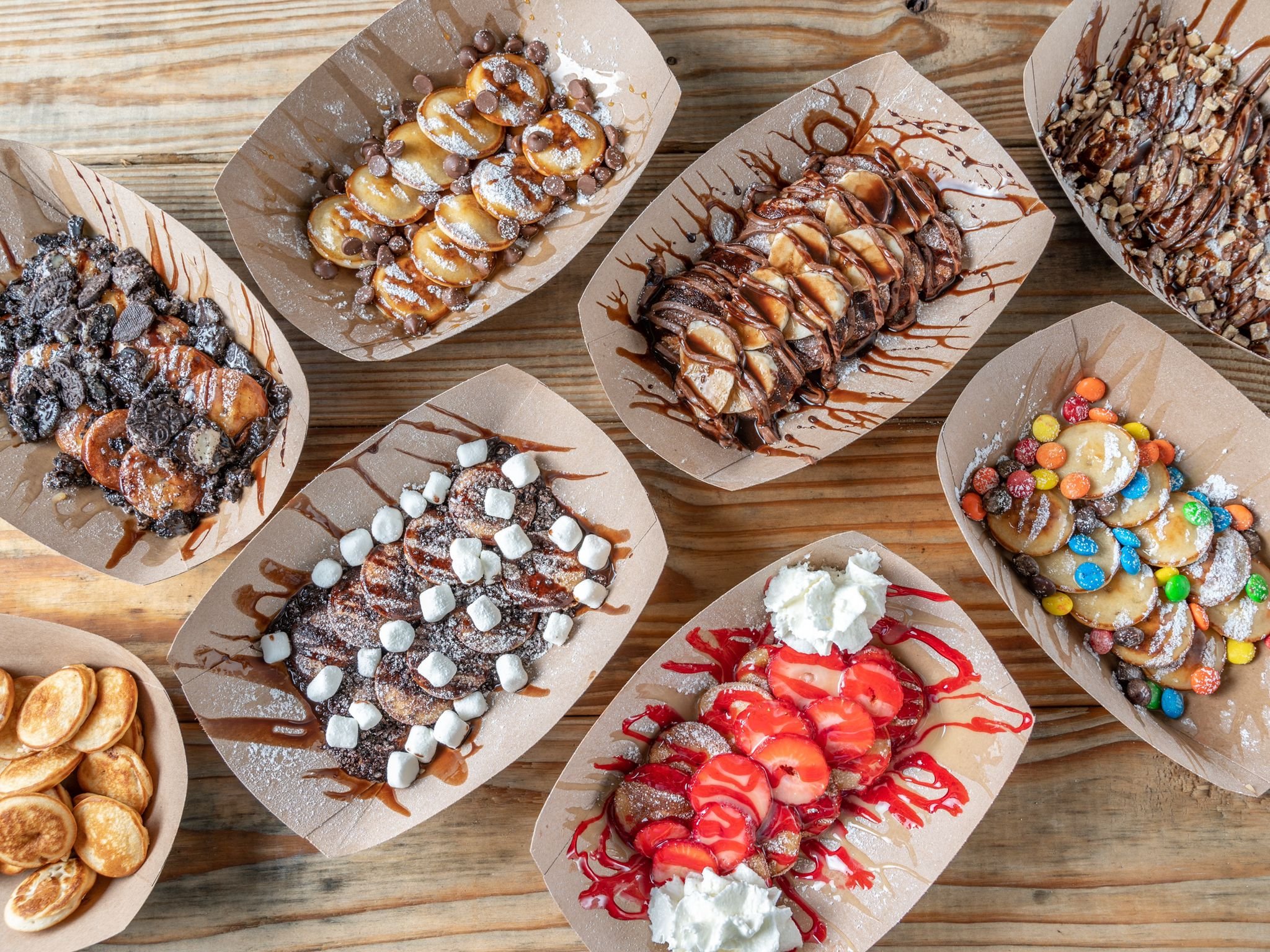 Assorted Belgian waffles topped with various toppings such as chocolate, marshmallows, whipped cream, strawberries, colorful candies, and sauces, arranged on a wooden table.