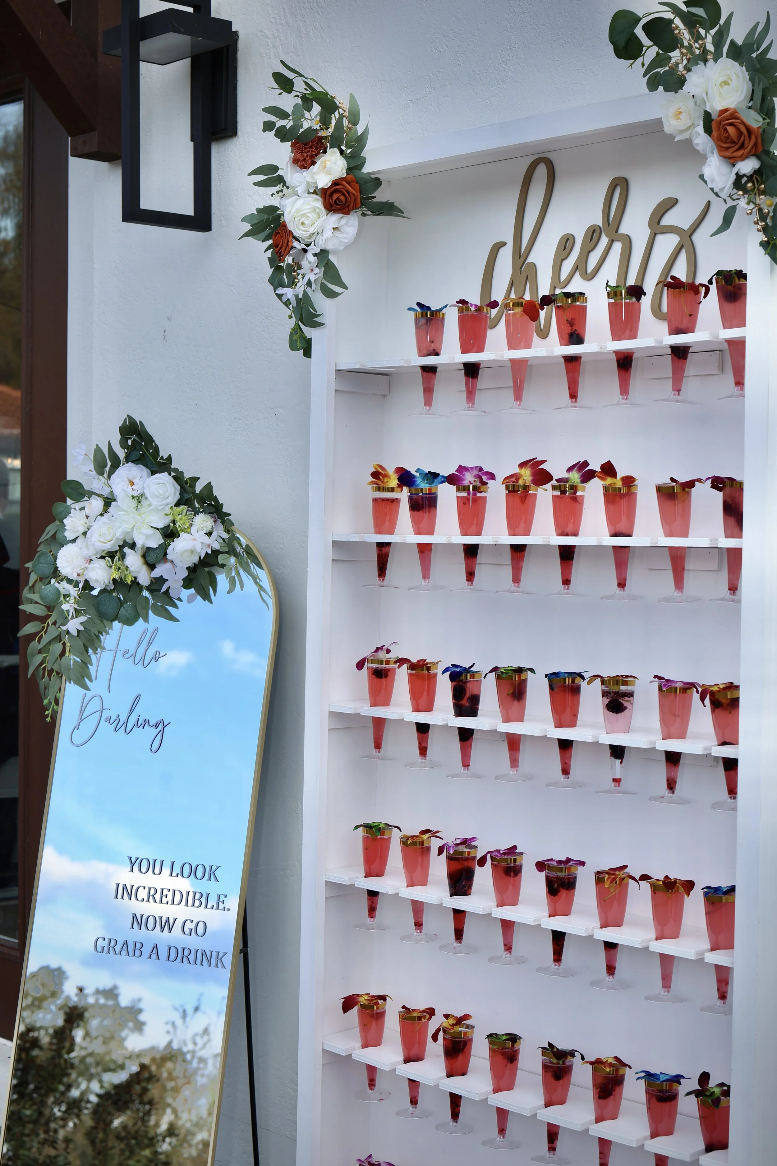A display of colorful beverages in glasses arranged on a white shelf with a 'Cheers' sign above, decorated with floral arrangements of white, rust, and green flowers. To the left, a tall mirror with flowers and a sign that says 'Hello Darling, You look incredible. Now go grab a drink'.