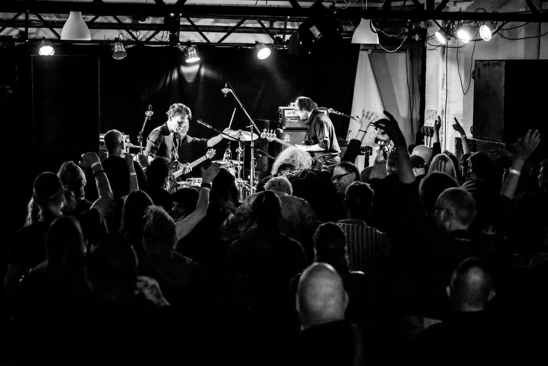 Black and white photo of a live music performance on stage with band members playing guitar and keyboard, audience members dancing and raising their hands in front of the stage in a concert setting.