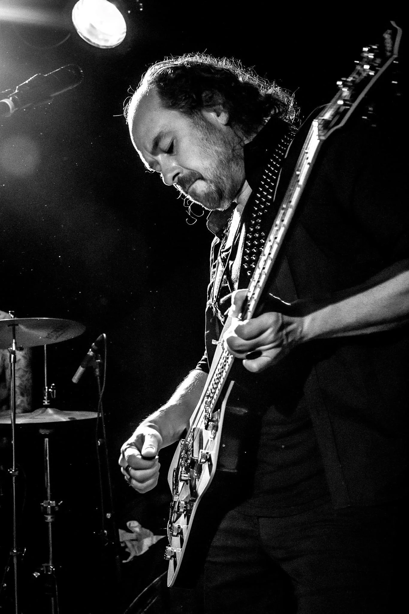 A male musician playing an electric guitar on stage, with a drum set visible in the background. The scene is lit with a spotlight, and the image is in black and white.
