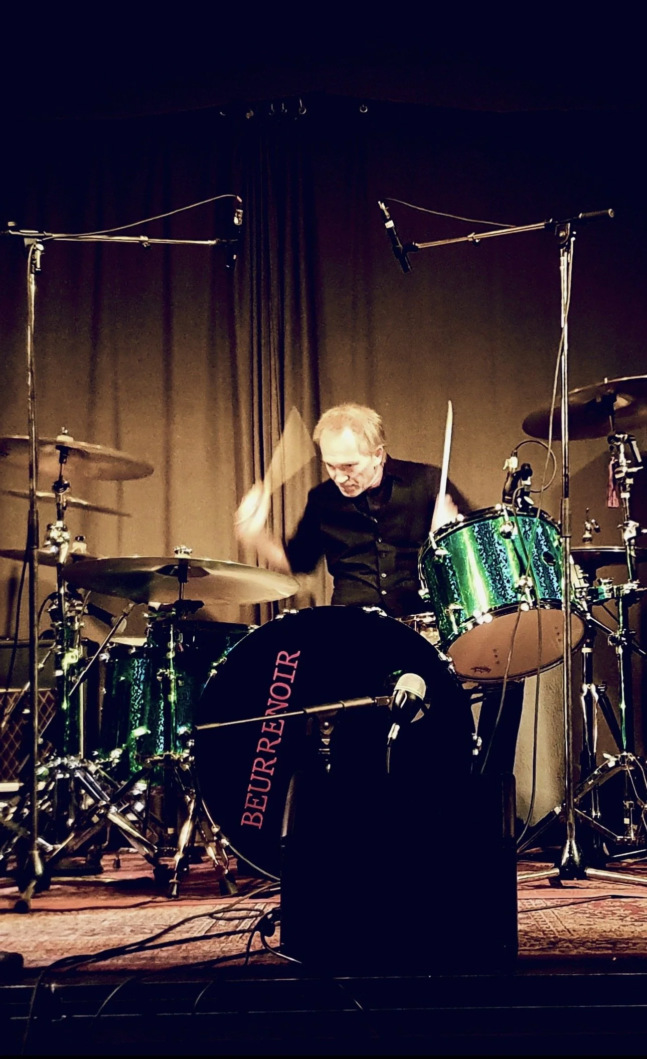 A drummer playing a green drum set on stage with a dark curtain backdrop, microphones above, and a black front bass drum with 'BEURRENOIR' written on it in pink.