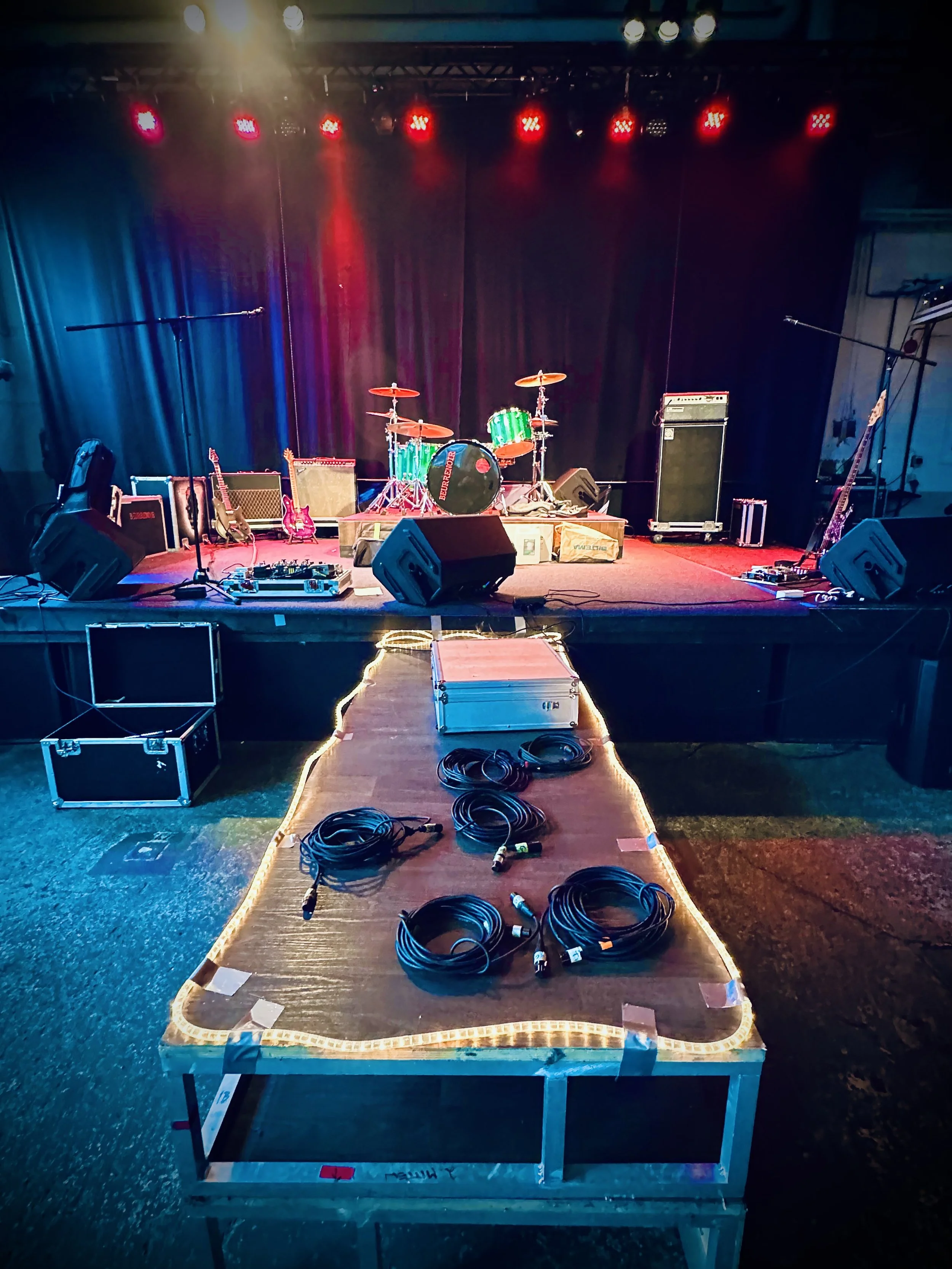 Stage with musical instruments including drums, guitars, and amplifiers, set up for a performance, with lighting and cables on the floor, and a table with coiled cables in the foreground.