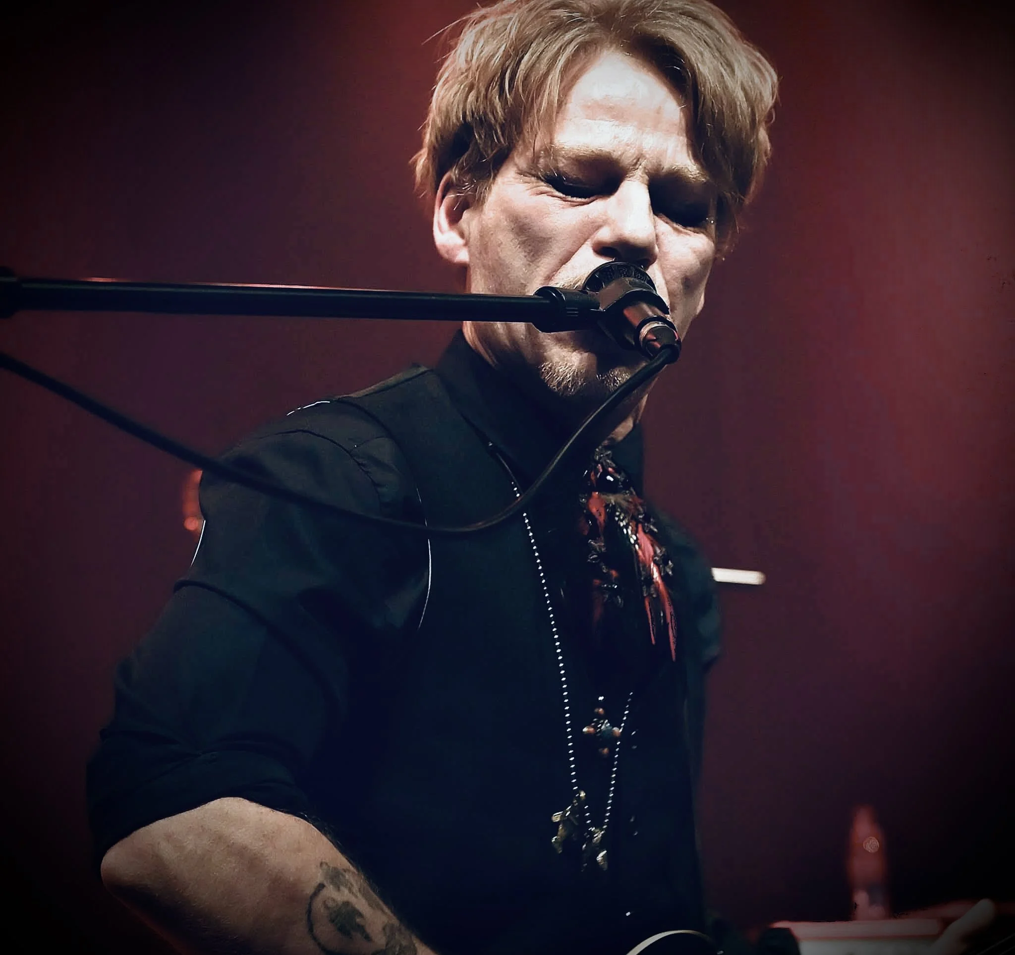 A musician singing into a microphone during a live performance, with a dark background and warm lighting.