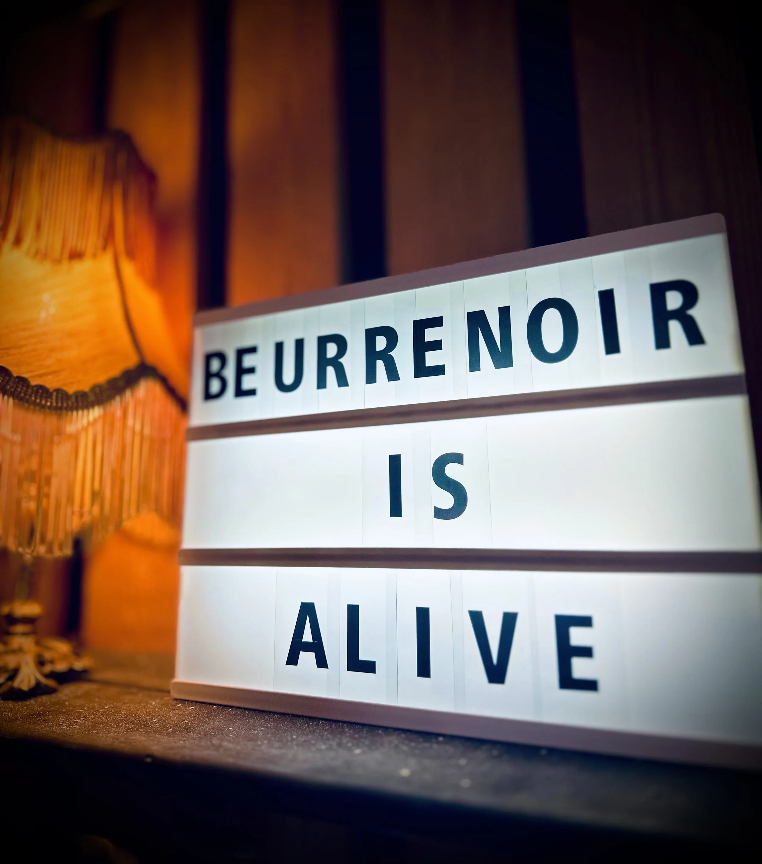 A lightbox sign displaying the words 'BEURRENOIR IS ALIVE' on a wooden surface, with a warm orange lamp and wooden panel background.