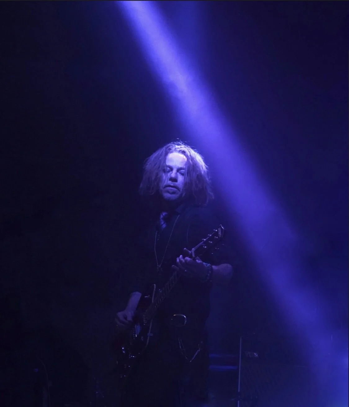 A male guitarist with shoulder-length hair performing on stage, illuminated by purple and blue stage lights, holding an electric guitar.