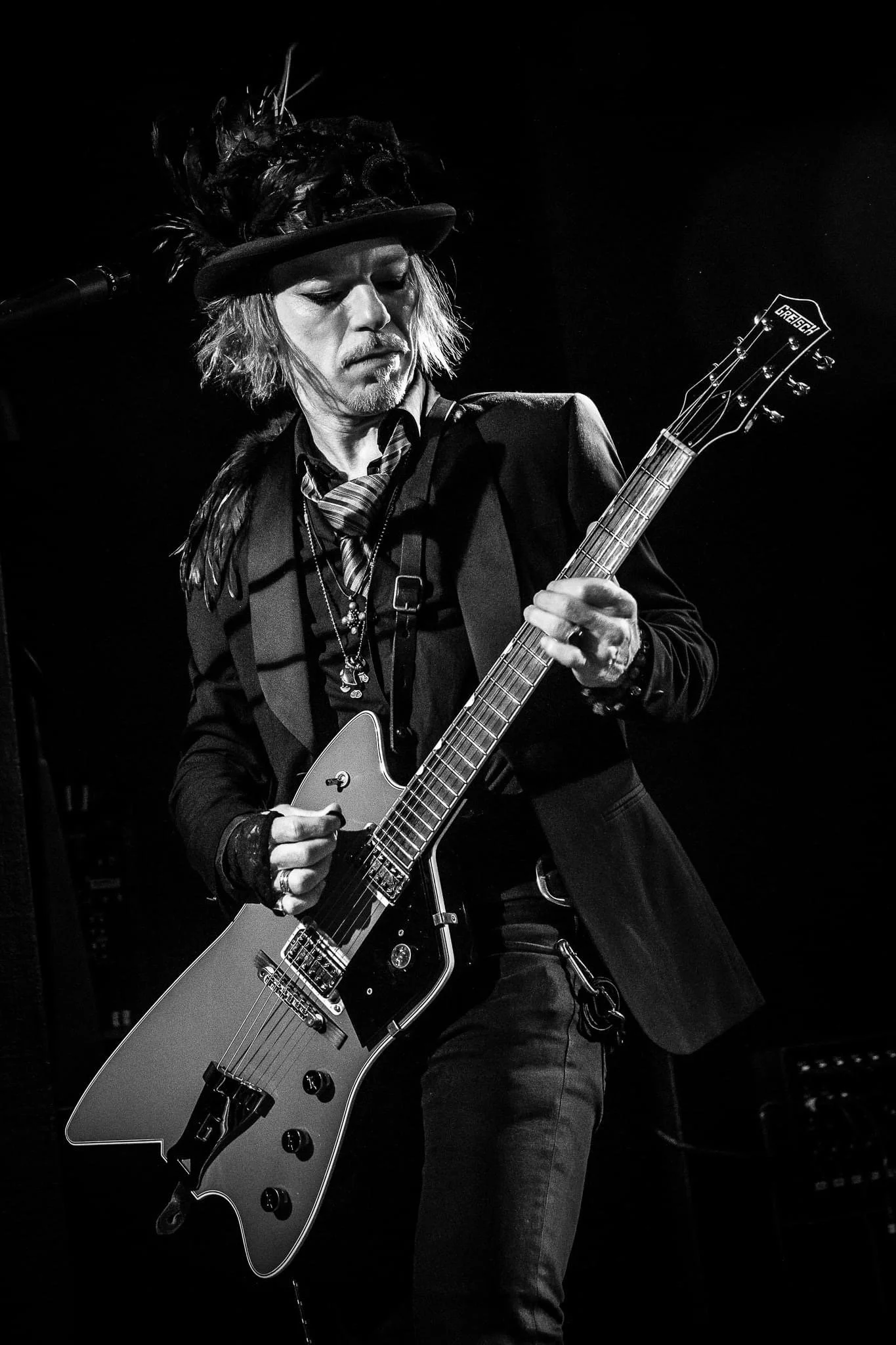 A man wearing a feathered hat and dark clothing playing an electric guitar on stage in black and white.