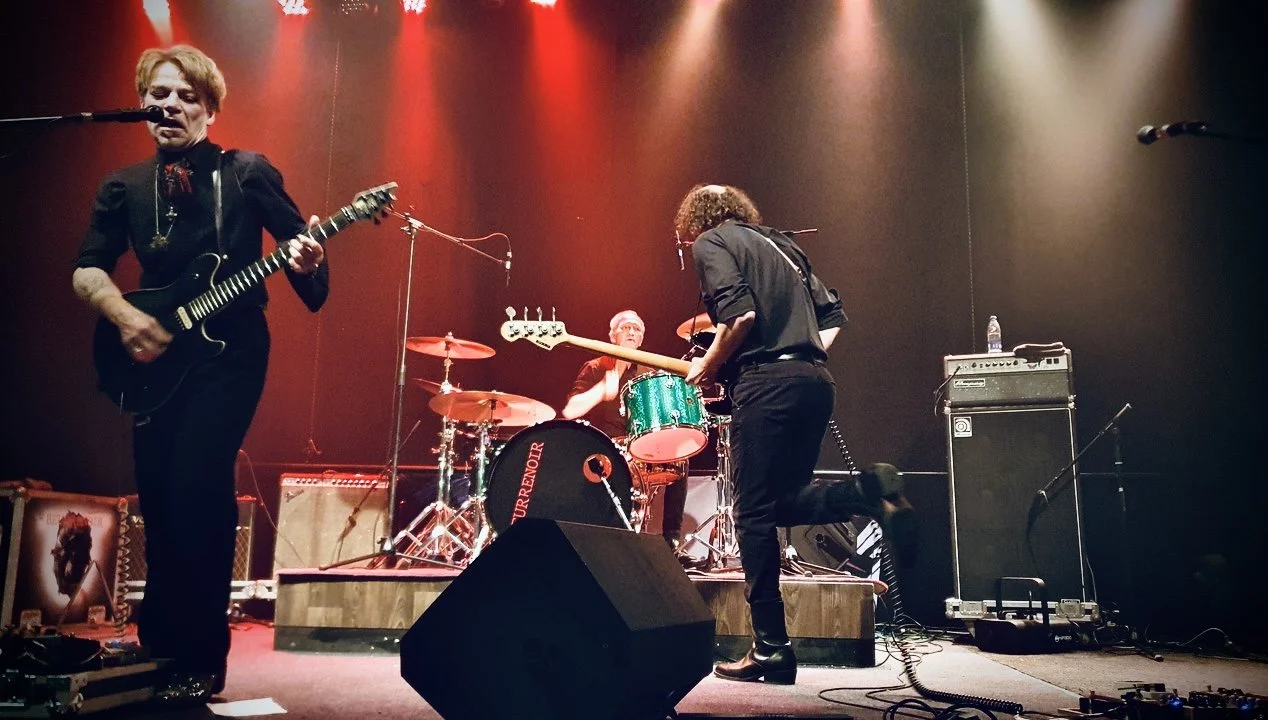 A band performing on stage with a guitarist singing into a microphone, a drummer playing drums, and a bass player kneeling while playing bass guitar, all under red stage lighting.